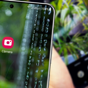 Mão segurando um smartphone Samsung apontado para um ambiente com luz natural, mostrando a tela com a interface da câmera.